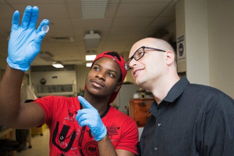 A student wearing blue gloves holds up and points to a sample while a professor investigates, as well.